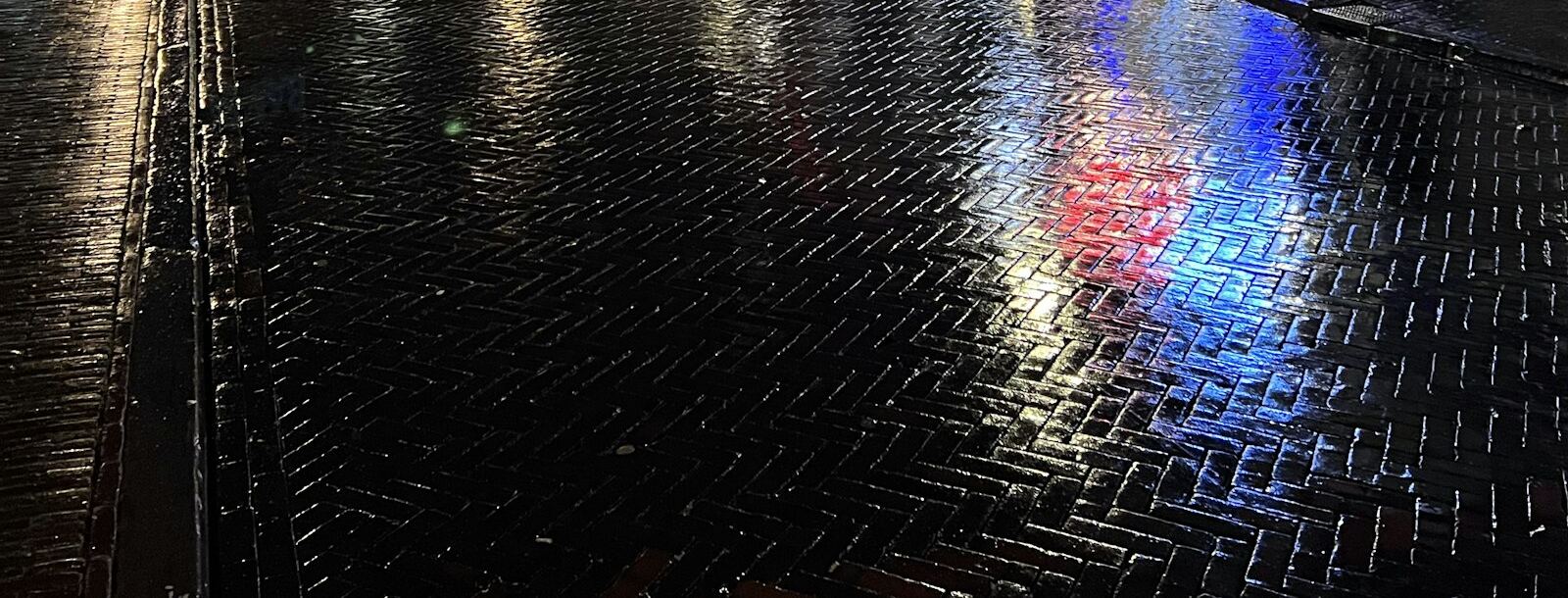 Natte klinkerstraat in een Nederlandse binnenstad op een regenavond, met de weerspiegeling van lantaarns en winkellicht
